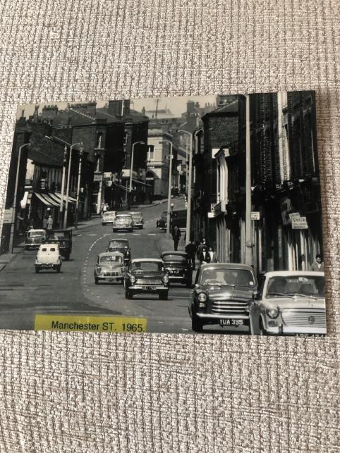 A black and white photo of a view down a busy road, which has plenty of cars on it. There are street lamps and houses lining the road, with a row of shops visible in the background. The road seems to curve off to the right, uphill from the photograph’s perspective. There is a yellow piece of paper stuck to the bottom, which reads Manchester ST. 1965