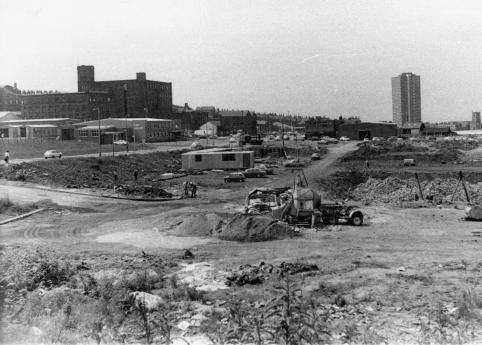A black and white photo showing clearance and construction work in the foreground. In the background you can see a couple of larger industrial buildings, and rows of terraced houses identifiable by their pointed roofs, as well as one tower block to the right of the frame.